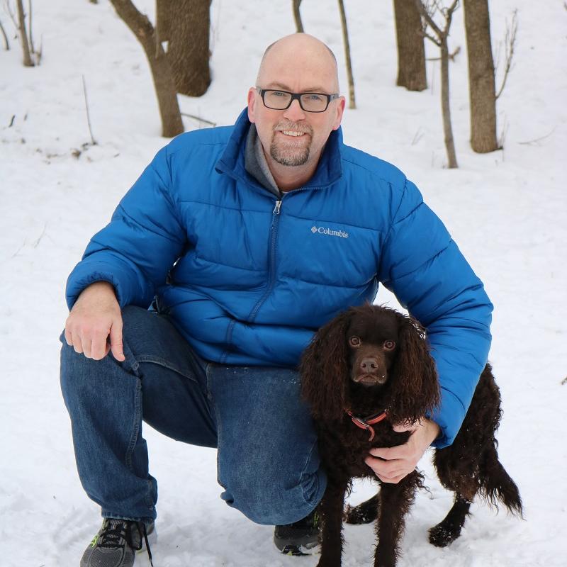 Man kneeling in the snow next to a brown dog