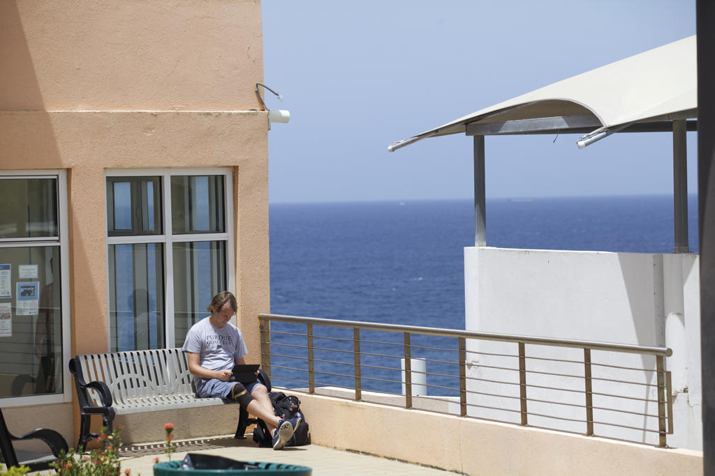 A man sits reading on a bench outside of a building, with the ocean in the background.