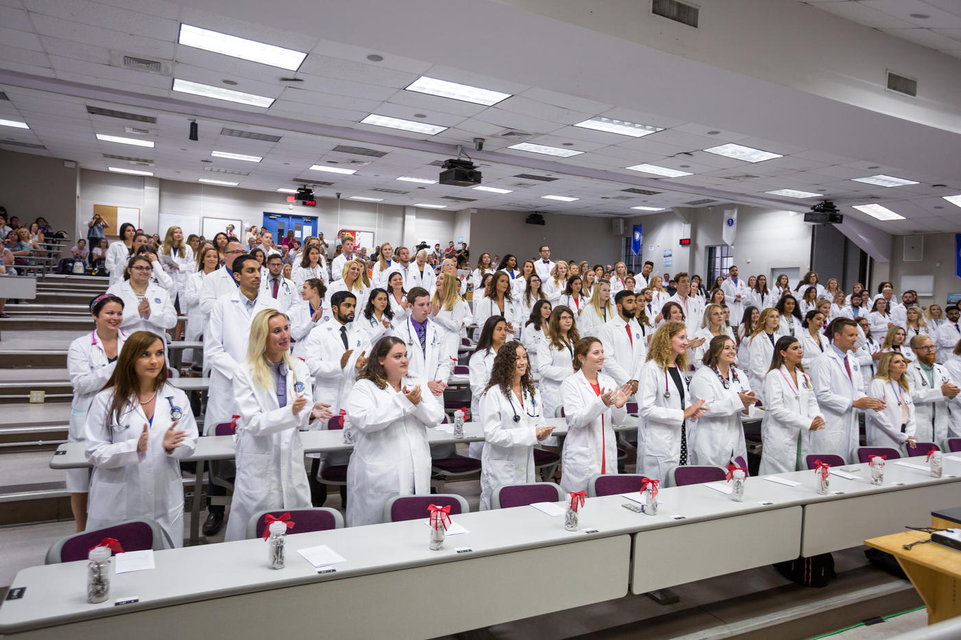 A classroom filled with students in lab coats, standing and applauding