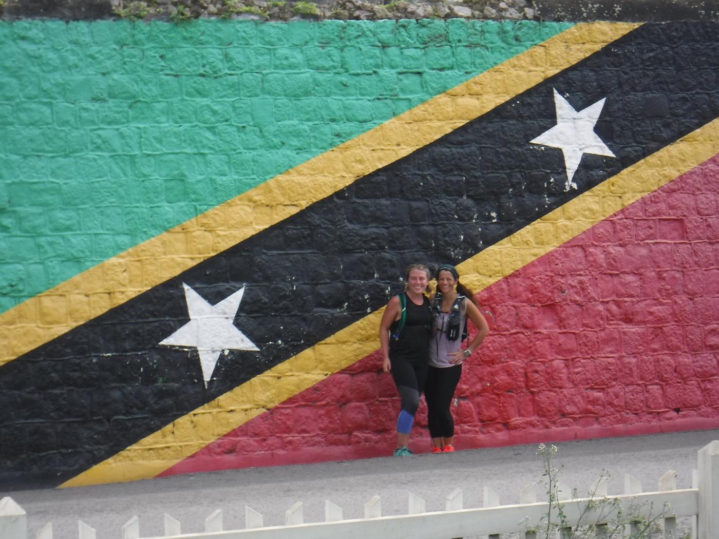 Two young women posing in front of a giant mural of the St. Kitts flag.